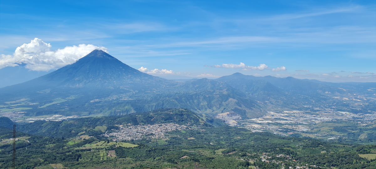A scenic landscape photograph of a large volcanic mountain under a bright blue sky with wispy clouds, overlooking a lush green valley and town.