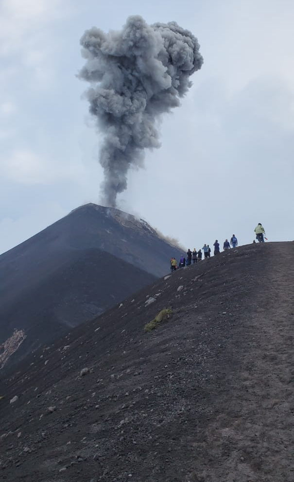 A volcanic eruption with a large gray ash plume rising into the sky, with a dark mountain slope in the foreground and a small group of people standing on the right side.