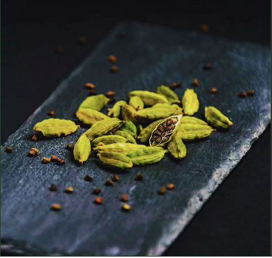 Green cardamom seeds with one cracked open seed showing the inner texture, scattered on a dark slate serving board against a black background.